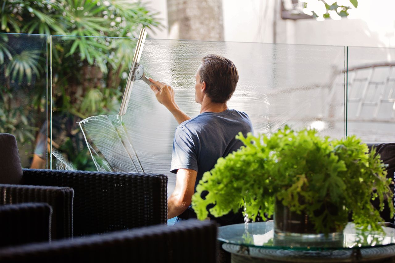services-01 A man outdoors is cleaning a glass barrier with a squeegee, with greenery all around.